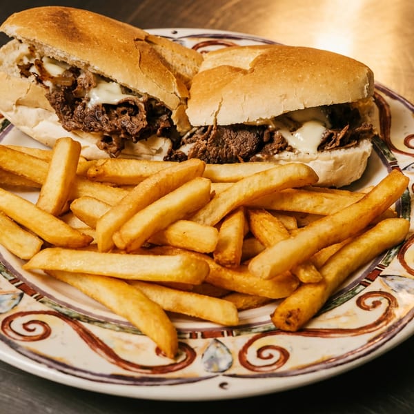 A cheesesteak sandwich cut in half, served with a pile of golden french fries on a decorative plate.