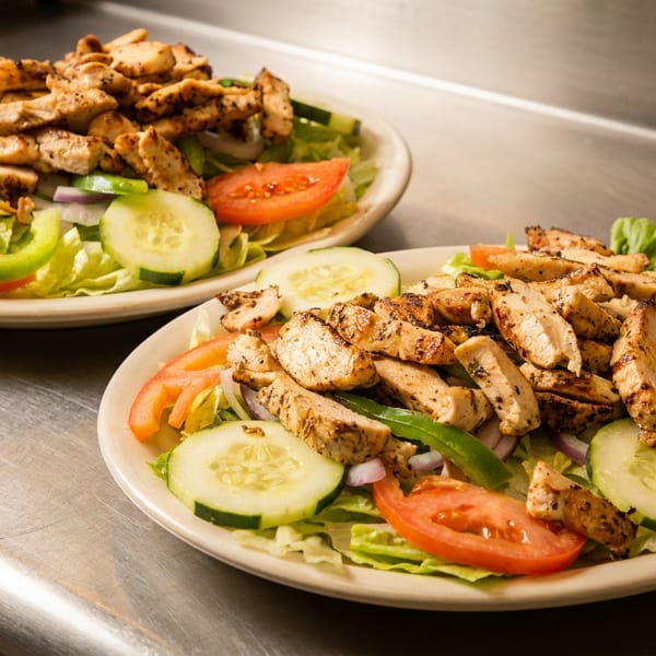 Two plates of grilled chicken salad with lettuce, cucumbers, tomatoes, and bell peppers on a metal counter.