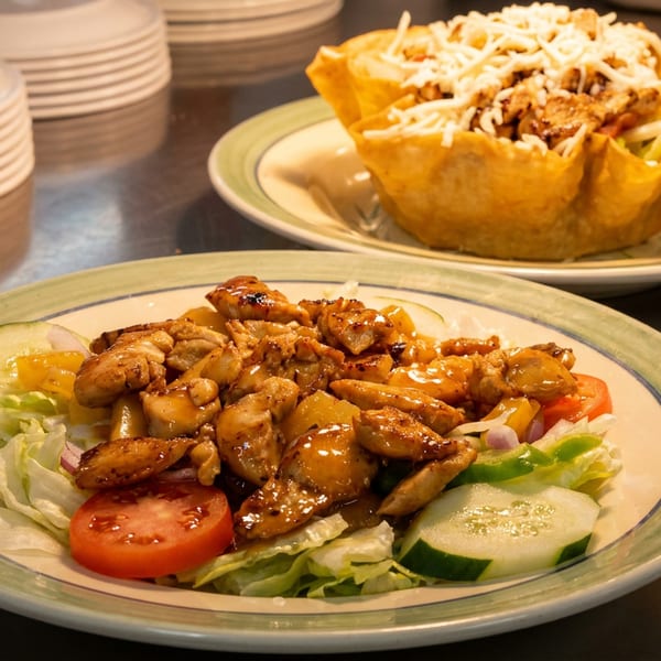 Two plates of food, one with chicken and vegetables, the other a taco salad in a fried shell.